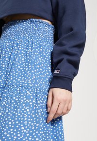 Sininen mekko, jossa on valkoisia pilkkuja ja rypytetty vyötärö, yhdistettynä tummansiniseen lyhythihaiseen collegepaitaan, jossa on pieni logo hihassa.
