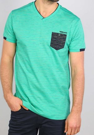 Mężczyzna ubrany w turkusowy T-shirt z dekoltem w serek w cienkie poziome paski oraz czarną kieszonką na piersi w paski, połączony z ciemnymi spodniami.