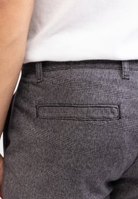 Grå rutiga byxor med en platt bakficka med en broderad kant, tillverkade av ett strukturerat vävt tyg.