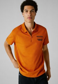 Oranžová polo košeľa z bavlny, s polovičným zipsom, vyšívaným logom na hrudi a krátkymi rukávmi, v kombinácii s tmavými džínsami.