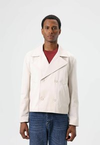 Lichtbeige dubbelgeknoopt jasje met een brede kraag, voorzien van twee voorzakken en knoopsluitingen, gedragen over een bordeauxrode top en spijkerbroek.