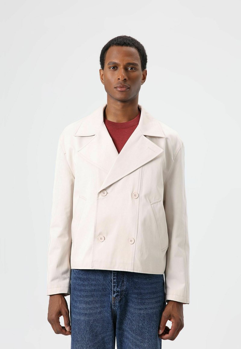 Lichtbeige dubbelgeknoopt jasje met een brede kraag, voorzien van twee voorzakken en knoopsluitingen, gedragen over een bordeauxrode top en spijkerbroek.