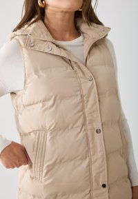 Gilet rembourré beige avec une fermeture éclair à l'avant et des boutons-pression. Présente une capuche, une poche latérale et une texture matelassée pour l'isolation.