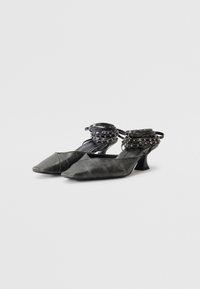 Chaussures noires à bout pointu avec une finition texturée, présentant un petit talon carré et des brides de cheville ornées.