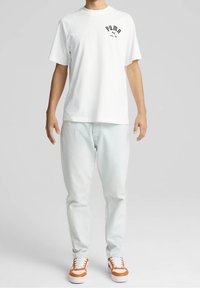 Camiseta blanca de manga corta con un logo negro de Puma en el lado izquierdo del pecho, combinada con jeans azul claro y zapatillas acentuadas en naranja.