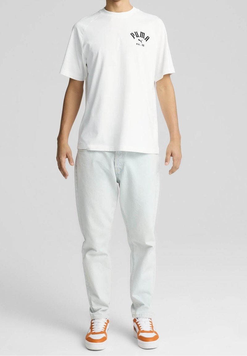 Camiseta blanca de manga corta con un logo negro de Puma en el lado izquierdo del pecho, combinada con jeans azul claro y zapatillas acentuadas en naranja.