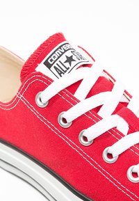 Sneaker in tela rossa con lacci bianchi. Presenta una suola in gomma, una striscia nera in contrasto e un'etichetta con la scritta "Converse All Star" sul linguetta.