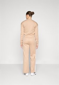 Fato de treino bege feito de tecido macio, com riscas laterais brancas, uma jaqueta cropped ajustada e calças longas descontraídas. Usado com tênis brancos.