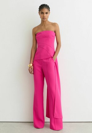 Fonott hajú nő élénk rózsaszín pánt nélküli felsőt és hozzá illő, bő szárú nadrágot visel, egyszínű fehér háttér előtt áll.