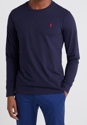 Navy long-sleeve t-shirt made of cotton with round neckline and embroidered red logo on the left chest, worn with blue pants.
