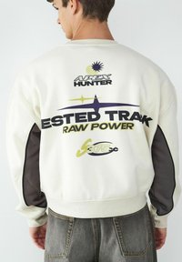 Biała bluza z kapturem z fioletowymi i zielonymi grafikami na plecach, z wyraźnym tekstem i kontrastującym ciemnoszarym panelem na rękawie.