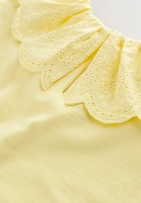 Žltý vrchný diel zo látky s oblým golierom s vyšívanými kvetinovými motívmi a očkovými výrezmi, ktoré vytvárajú textúrovaný vizuálny kontrast.