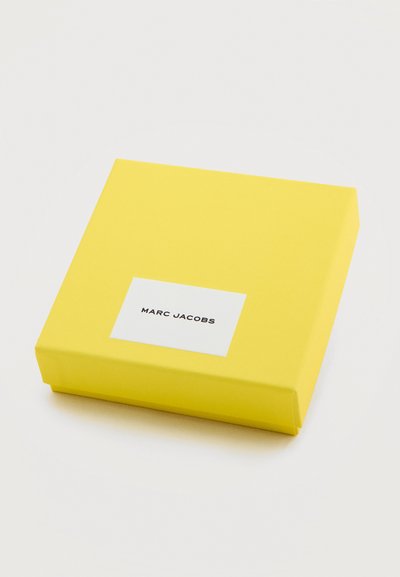 Κίτρινο τετράγωνο κουτί με λευκή ετικέτα στο κέντρο που εμφανίζει το "MARC JACOBS" με μαύρα κεφαλαία γράμματα.