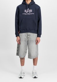 Uomo che indossa un hoodie navy di Alpha Industries, pantaloni corti di jeans grigi chiari, calze bianche e scarpe da ginnastica bianche, in piedi davanti a uno sfondo neutro.