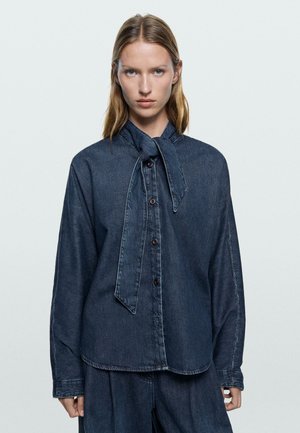 Vrouw die een donker denim overhemd met knopen en lange mouwen draagt, met een geknoopt sjaaltje om de hals, gecombineerd met bijpassende denim broek, tegen een effen achtergrond.