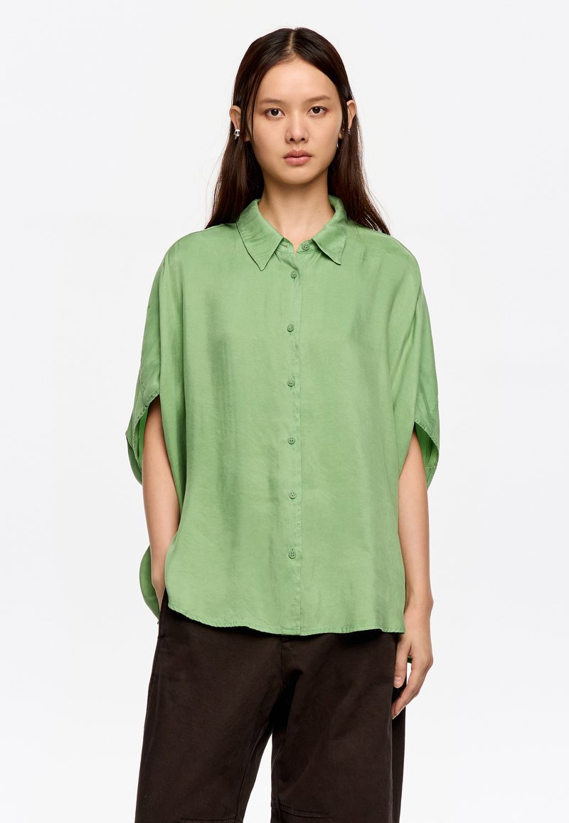 Camisa de botones verde claro, de manga corta, ajuste relajado, hecha de una tela suave. Combinada con pantalones oscuros, el modelo se encuentra de pie frente a un fondo blanco.