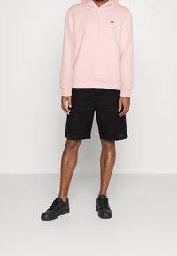 Sudadera con capucha de color rosa claro, con un bolsillo canguro en la parte frontal, combinada con pantalones cortos negros y zapatillas negras. Diseño simple y material de algodón.