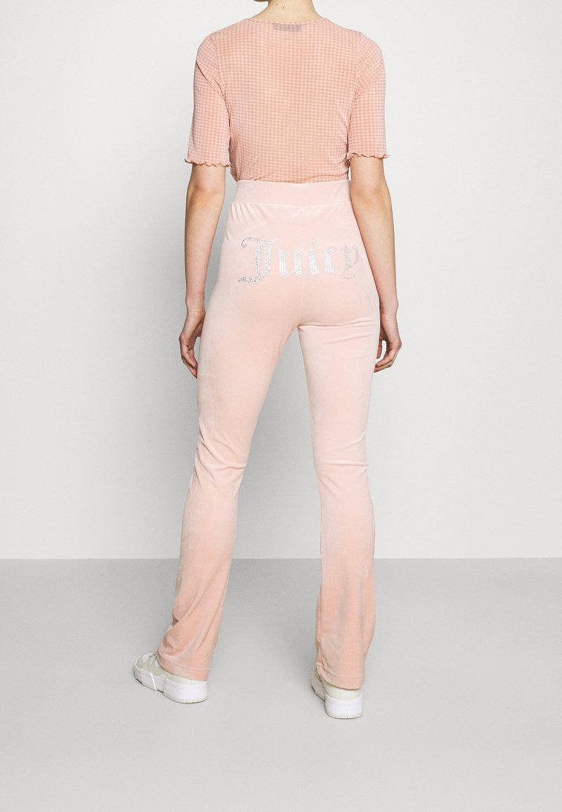 Personne portant un pantalon en velours rose clair avec "Juicy" en strass à l'arrière, accompagné d'un haut à carreaux roses et de baskets blanches.