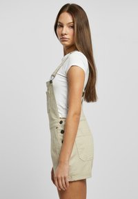 Tânără cu păr lung și castaniu, purtând un tricou alb și un salopetă scurtă bej, stând pe partea laterală, pe un fundal simplu.