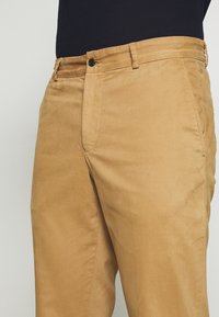 Chinos kaki en coton avec une texture lisse. Comprend une fermeture à bouton, des passants de ceinture et des poches latérales.