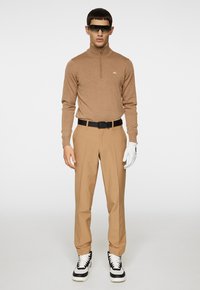 Brun zip-up tröja, tan byxor, svart bälte, vit handske på vänster hand och sneakers med svart och vita accenter, modell poserar mot en vit bakgrund.