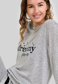 Mujer sonriente con cabello rizado marrón que lleva un sudadera gris jaspeada con el texto "soñadora para siempre" en negro en el frente.
