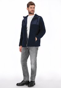 Chaqueta de forro polar azul marino con capucha, que presenta un panel contrastante azul marino en el pecho. Combinada con vaqueros grises y botas negras.