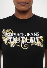 Juodas medvilnės marškinėliai su išraiškinga auksine ir balta siuvinėjimo ištrauka, kurioje yra užrašas "VERSACE JEANS COUTURE" dekoratyviame dizaine.