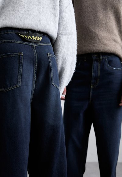 Dark blue jeans with yellow stitching and pockets, featuring a visible label at the back. Sweater textures are soft and light gray.