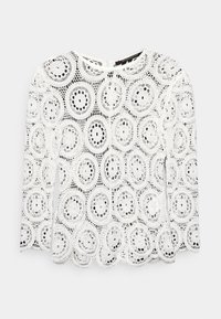 Blouse en dentelle blanche à manches longues, présentant des motifs circulaires et un ourlet festonné. Matériau léger et transparent avec un col rond.