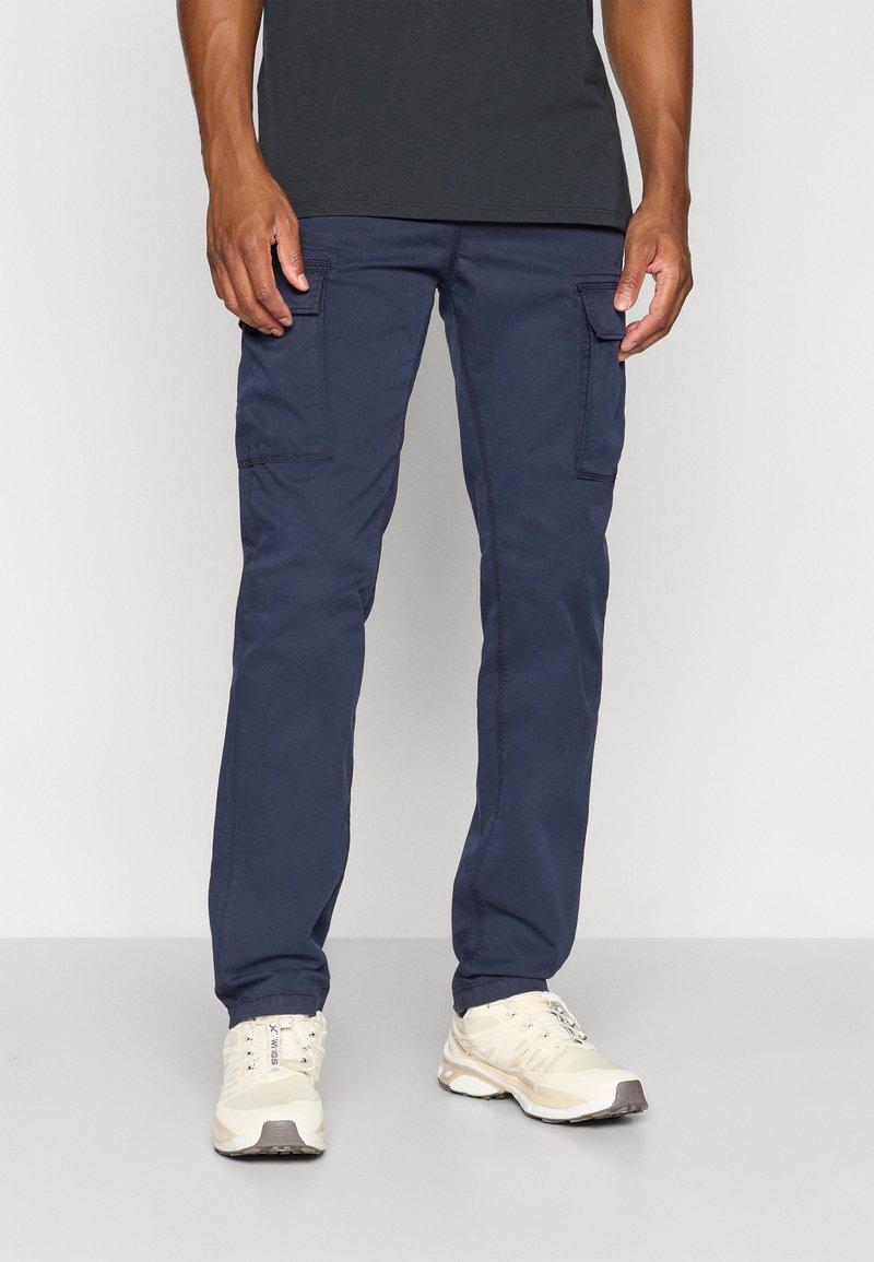 Pantalons cargo navy à coupe slim, fabriqués en un mélange de coton résistant, dotés de poches latérales et de détails de couture subtils. Associés à des chaussures de sport légères.