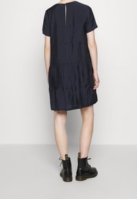 Person, die ein kurzärmeliges navy-blaues Kleid mit gerafftem Rockdetail trägt, kombiniert mit schwarzen Schnürstiefeln, steht vor einem schlichten Hintergrund.