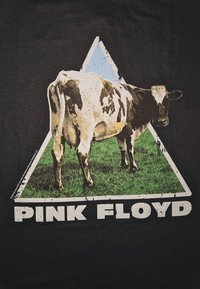 Svart t-skjorte med grafikk av en brun og hvit ku innenfor et blått og grønt trekantet design, med "PINK FLOYD" trykket under i fete hvite bokstaver.