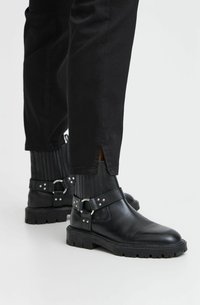 Juodos odinės kulkšninių botai su stambiu padu, šonine elastine juostele ir reguliuojama dirželiu su metaliniu užsegimu. Lygus padas ir tekstūruotas paviršius.