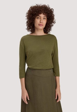 Donna con capelli ricci che indossa un top a maniche lunghe verde oliva e una gonna abbinata, in piedi contro uno sfondo chiaro e uniforme.