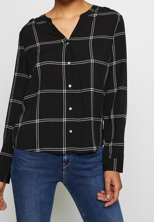 Kvinde iført en sort skjorte med hvide tern og blå jeans, vist fra nakken til hofterne.