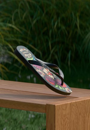 Fekete pántos, színes virágmintás flip-flop szandál egy faasztal szélén egyensúlyoz a szabadban.