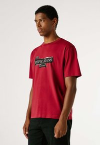 Camiseta roja de algodón con mangas cortas, cuello redondo y un gráfico con etiqueta negra en el frente, combinada con pantalones negros.