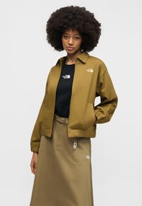 Γυναίκα με σγουρά μαλλιά που φοράει καφέ σακάκι με φερμουάρ, μαύρο T-shirt και καφέ φούστα που ταιριάζουν, όλα με λογότυπα The North Face.