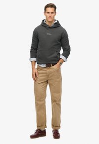 Sudadera gris oscura con bolsillo tipo canguro, logotipo bordado, superpuesta sobre una camisa de cuello azul claro. Pantalones color beige y zapatos náuticos granate completan el look.