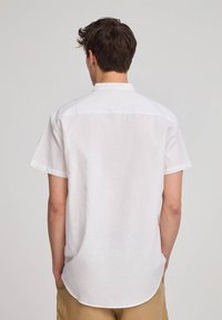 Camicia bianca a maniche corte in lino leggero, con colletto standard e orlo curvo, caratterizzata da una trama sottile. Vista da dietro.