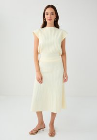 Abito plissettato giallo crema con maniche a cappuccio e vita aderente, abbinato a sandali con tacco color nocciola. Tessuto morbido e design fluido.