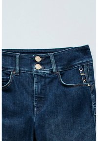 Jeans de mezclilla azul oscuro con dos botones de latón, bolsillos frontales y un detalle de cadena en el lateral. La tela tiene una textura suave y un ligero desgaste.