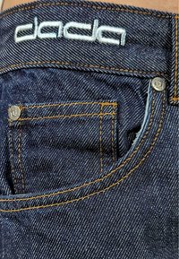 Dark denim fabric with orange stitching. Features a small front pocket, metal hardware, and "dada" embroidered logo in light blue.