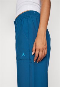 Teal sportovní kalhoty s elastickým pasem, které mají boční kapsu a malou modrou nášivku Jumpman na látce.