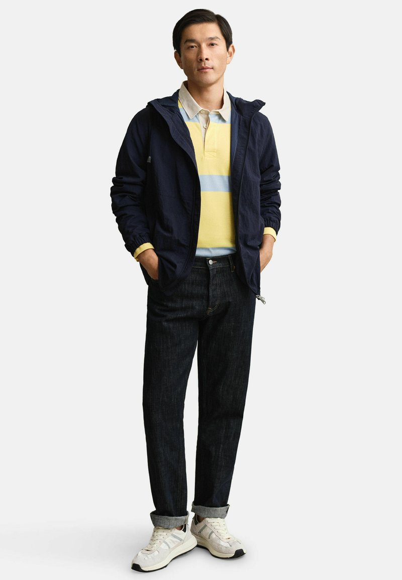 Homme debout les mains dans les poches, portant une veste zippée bleu marine, un polo rayé jaune et bleu, un jean foncé et des baskets blanches.