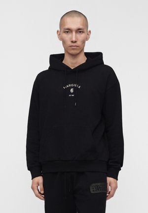 Homme au crâne rasé portant un sweat à capuche noir avec le texte blanc "MARGIELA 6 EST 1997", debout devant un fond clair uni.
