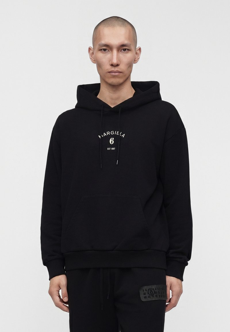 Man met geschoren hoofd die een zwarte hoodie draagt met witte tekst "MARGIELA 6 EST 1997", staand tegen een effen lichte achtergrond.