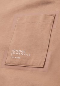 Beigenvärinen kangaspaita, jossa on neliönmuotoinen rintatasku ja valkoinen teksti "OMBRE STREET STYLE since 1985" taskussa.