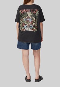Zwarte oversized t-shirt met een kleurrijk graphic ontwerp van tropische elementen, tekst en bloemen op de achterkant, gecombineerd met spijkerbroeken.